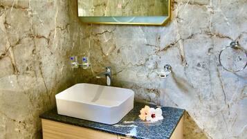 A bathroom with a marble wall, a white sink, a wooden vanity, a mirror, and a trash can.