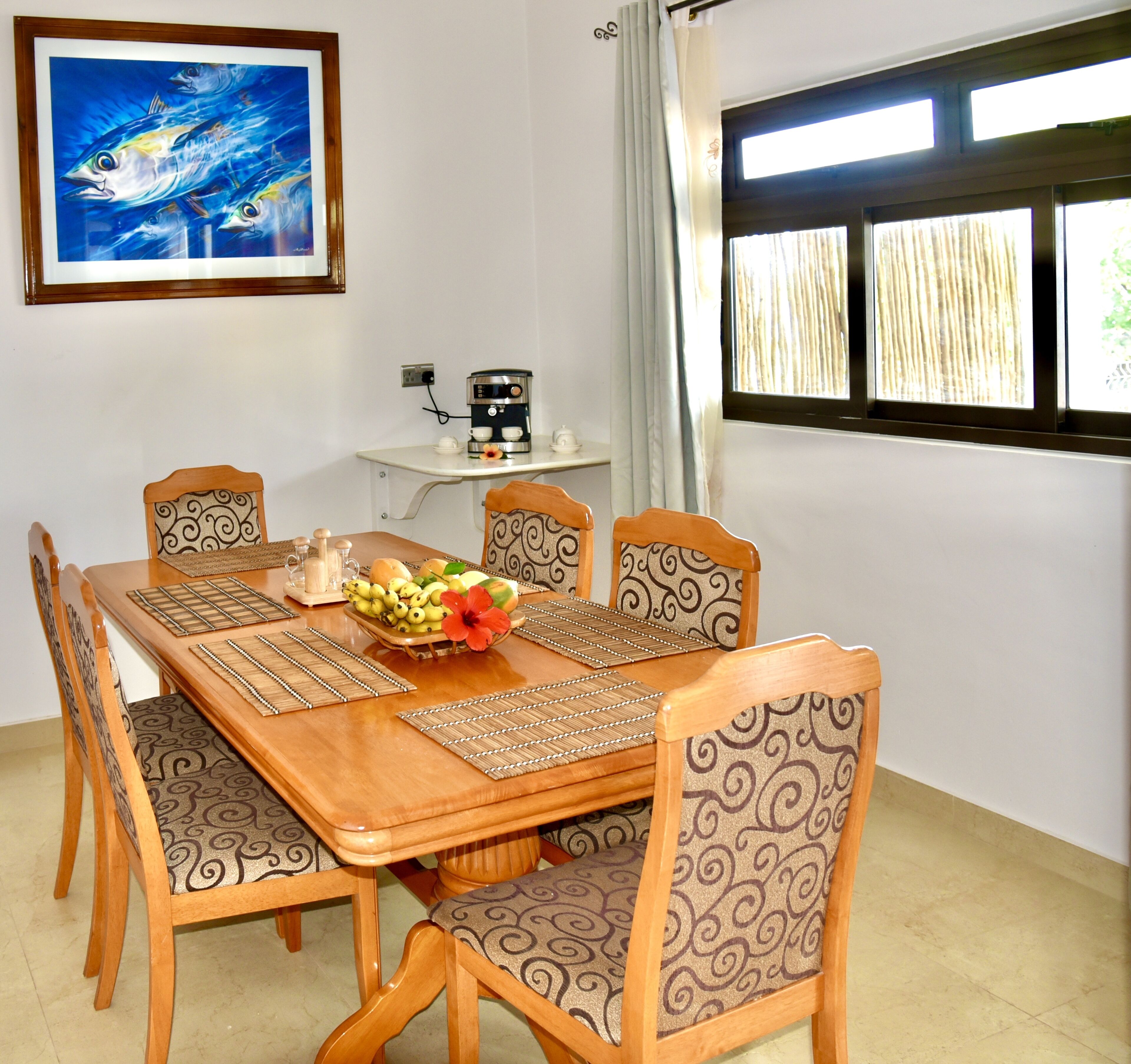 A dining area with a wooden table, four chairs, a coffee machine, and a fruit bowl.