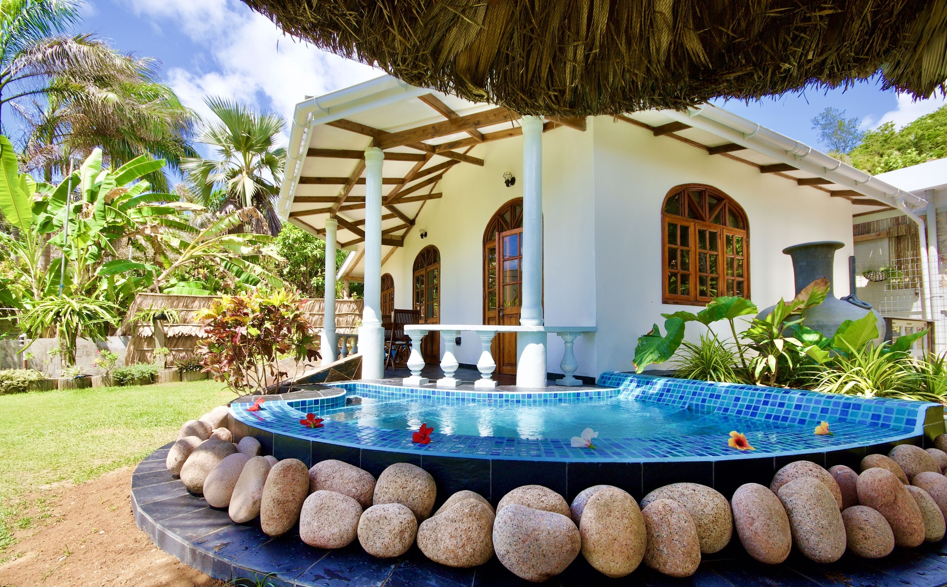 Un espace piscine agrémenté d’une structure à toit de chaume, d’une bordure en pierre et d’un jardin aux allures tropicales.