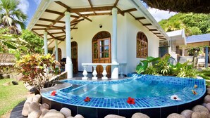 A pool with a tiled edge, surrounded by rocks, and a building with a thatched roof and columns. - Sea Splash & La Petite Maison (Praslin Island)