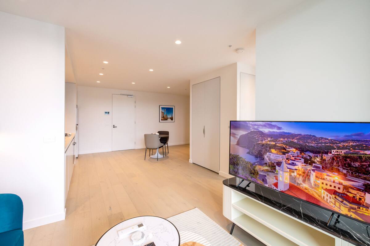 A modern living room with a flat-screen TV, a dining area with a table and chairs, and a view of a coastal town.