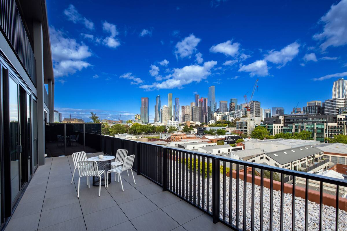 A balcony with white chairs and a table, overlooking a city skyline with modern buildings and a clear blue sky.