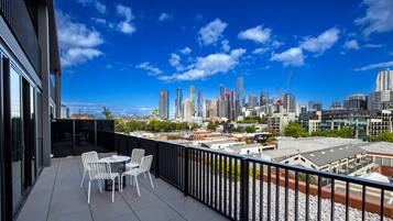 A balcony with white chairs and a table, overlooking a city skyline with modern buildings and a clear blue sky.