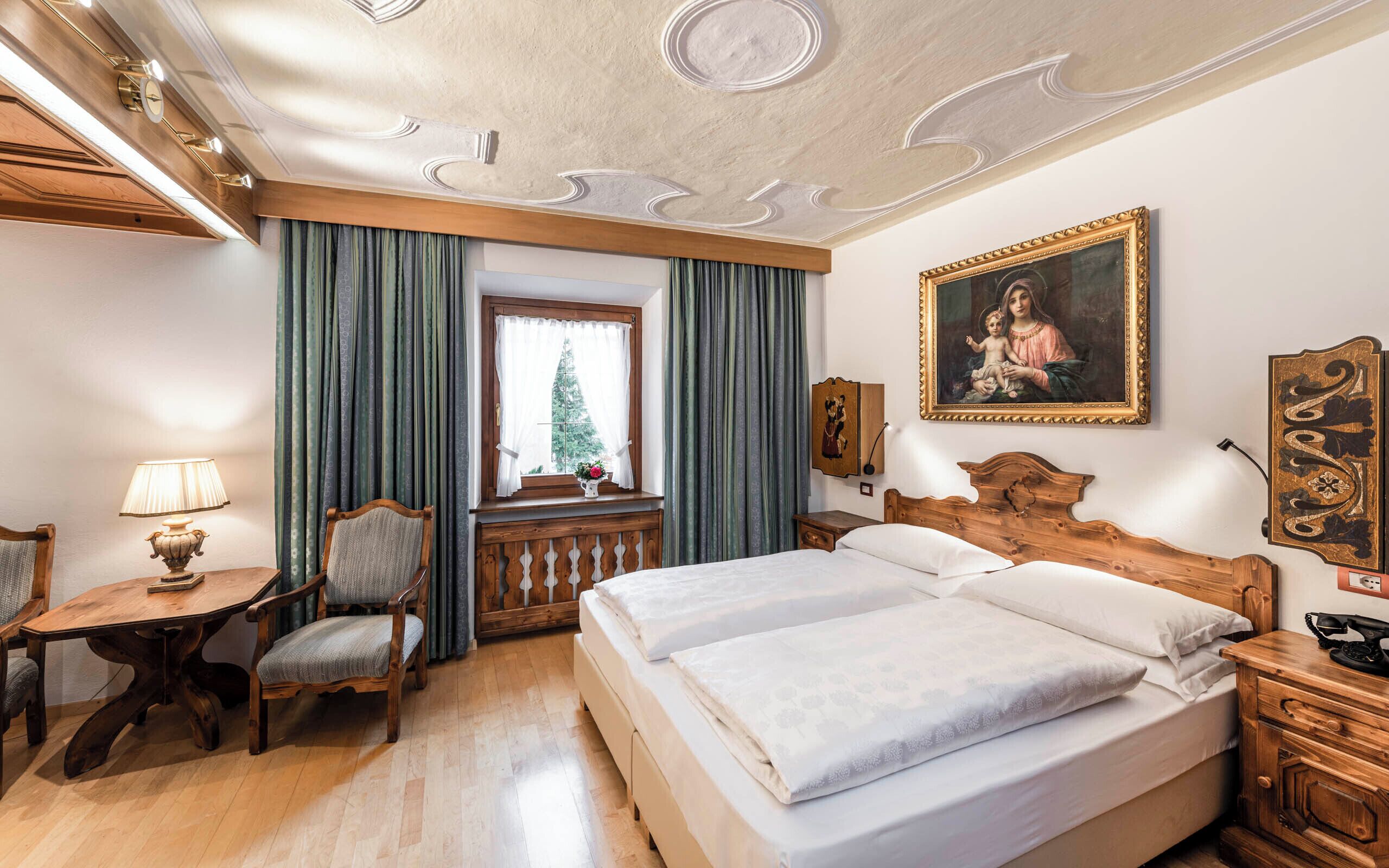 A hotel room with a wooden bed, a desk with a lamp, a chair, and a window with curtains.