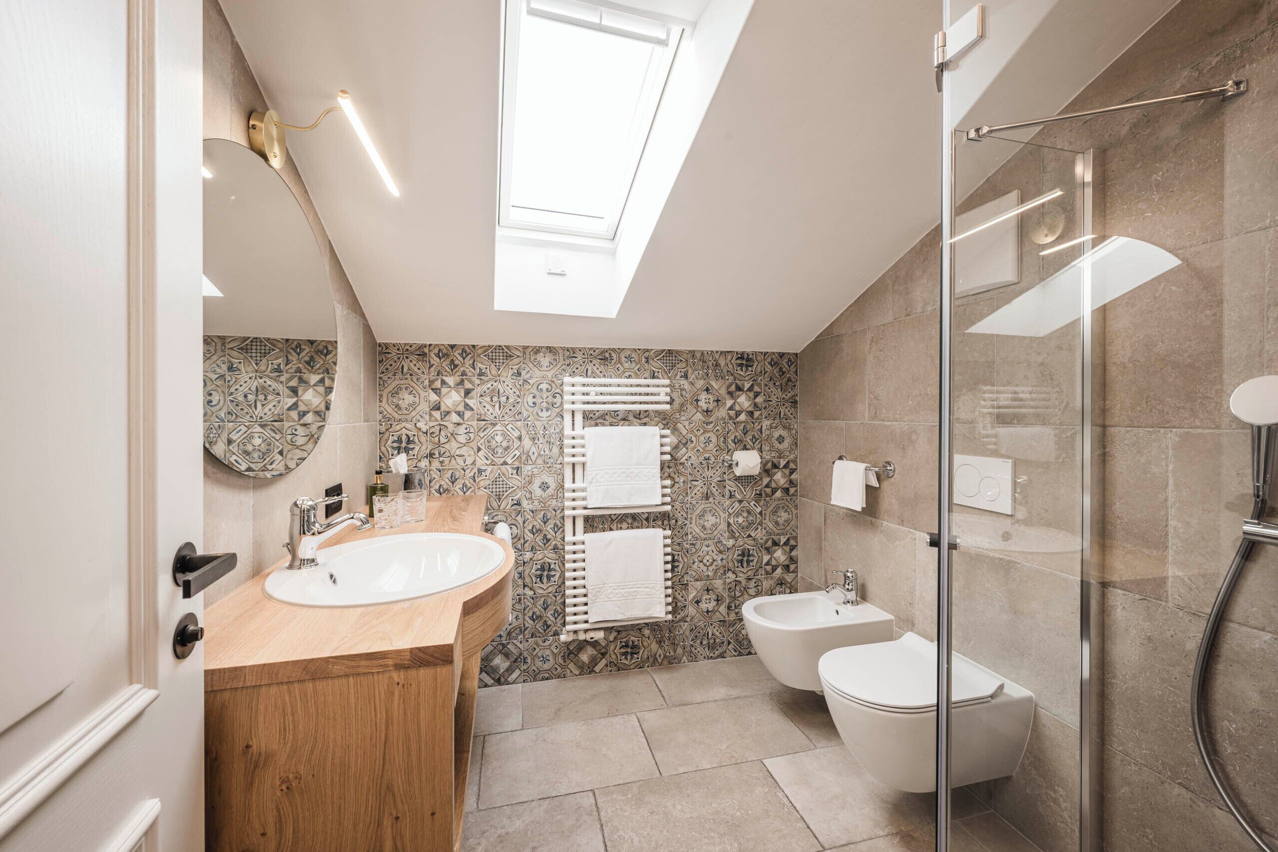 A modern bathroom with a skylight, a wooden vanity, a freestanding bathtub, and a glass-enclosed shower.
