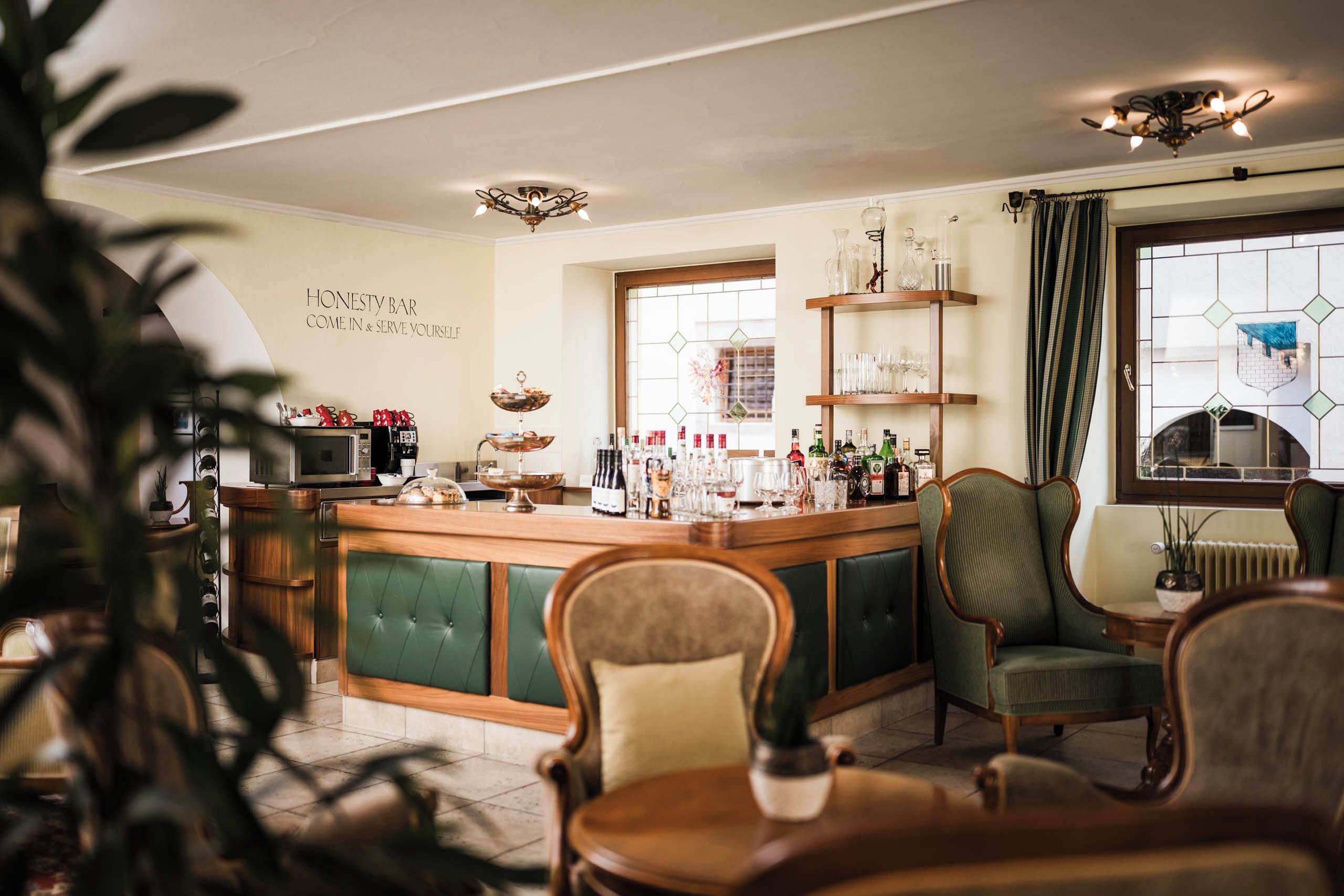 A bar area with a green cushioned bar counter, shelves with glasses and bottles, and a sign that reads 'HONESTY BAR COME IN & SERVE YOURSELF'.