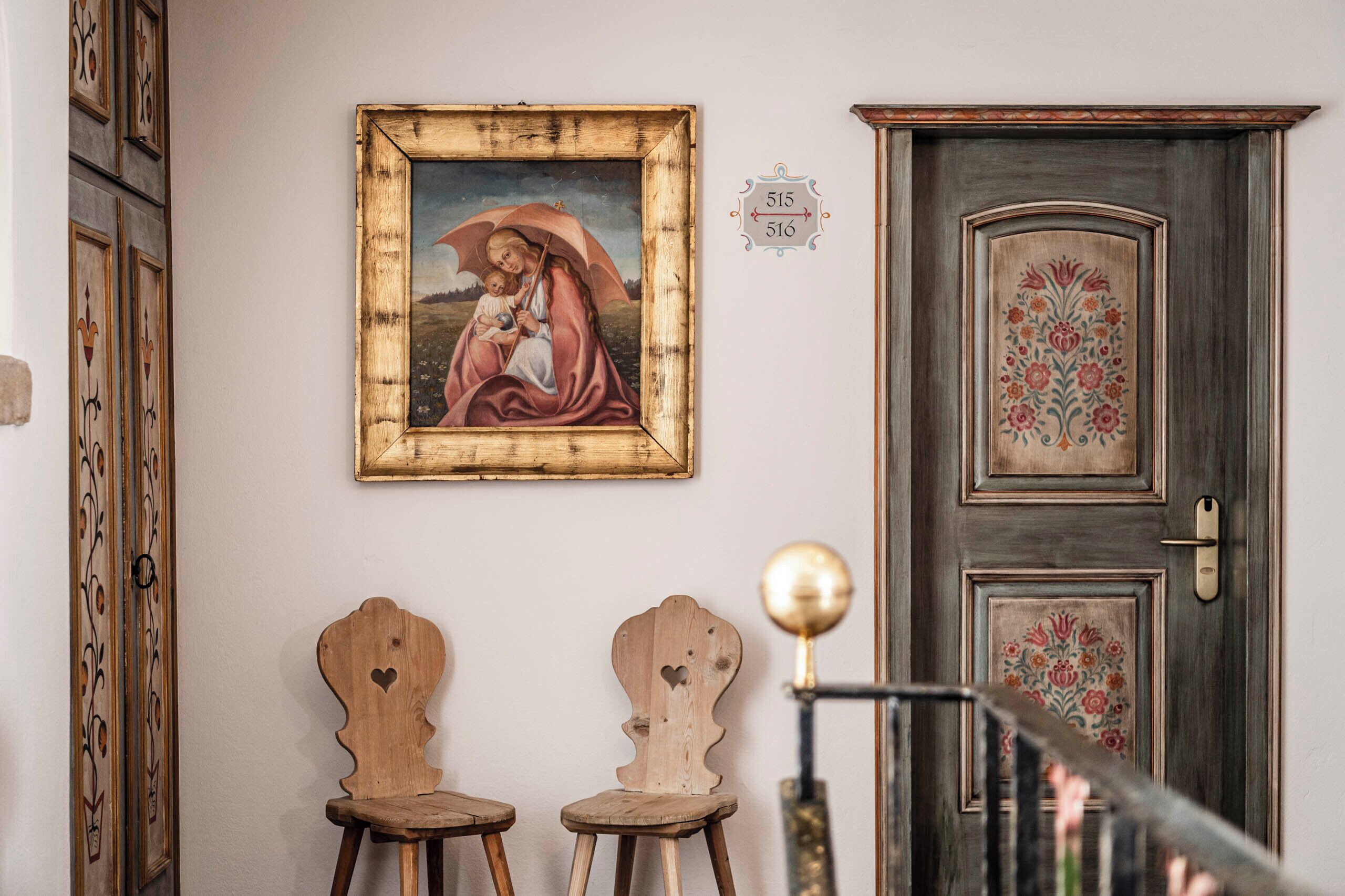 Una habitación con un cuadro enmarcado, dos sillas de madera, una puerta con motivos florales y una placa numérica colocada en la pared.