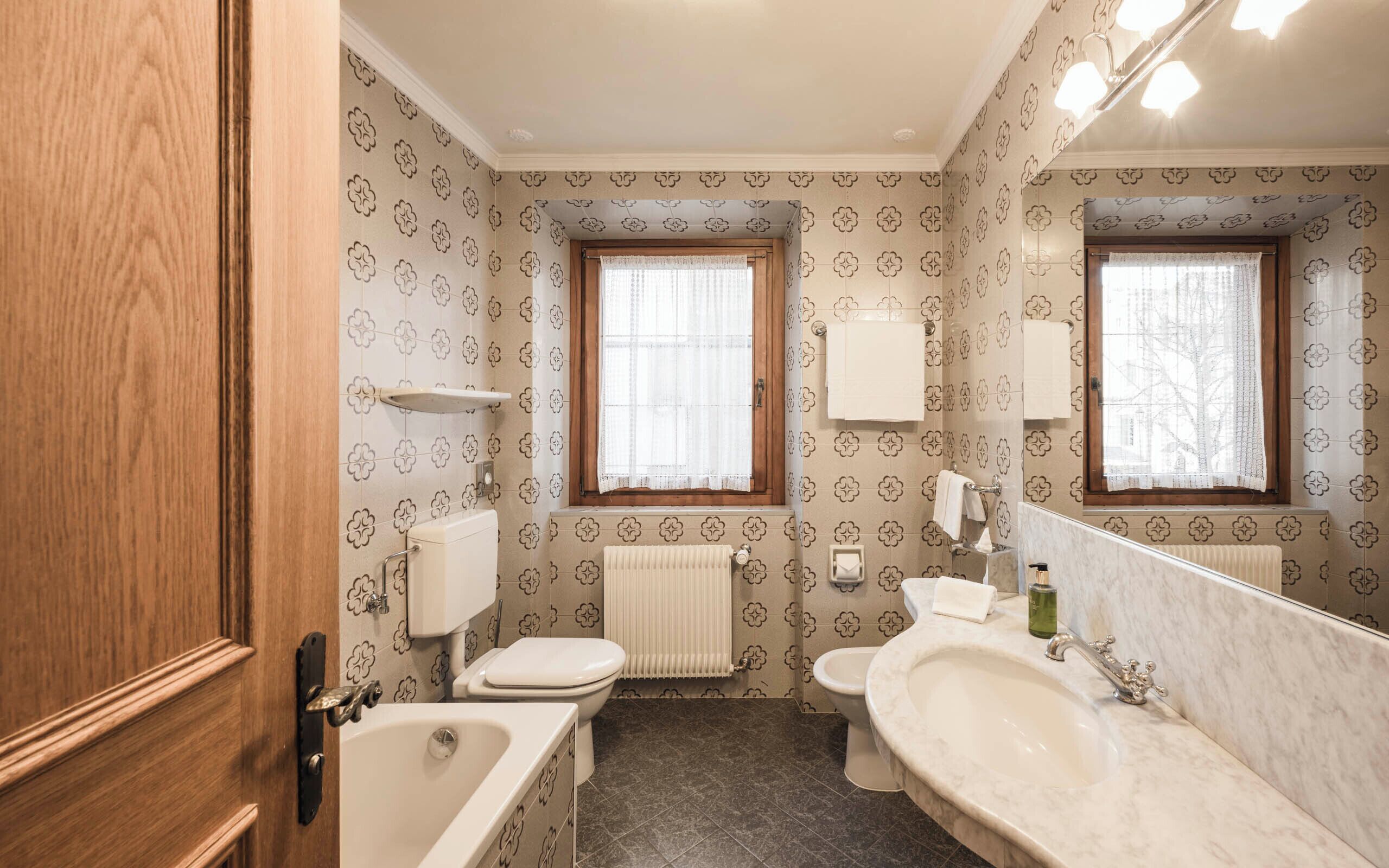 A bathroom with a toilet, sink, and bathtub. There are two windows with curtains, a towel rack, and a mirror.
