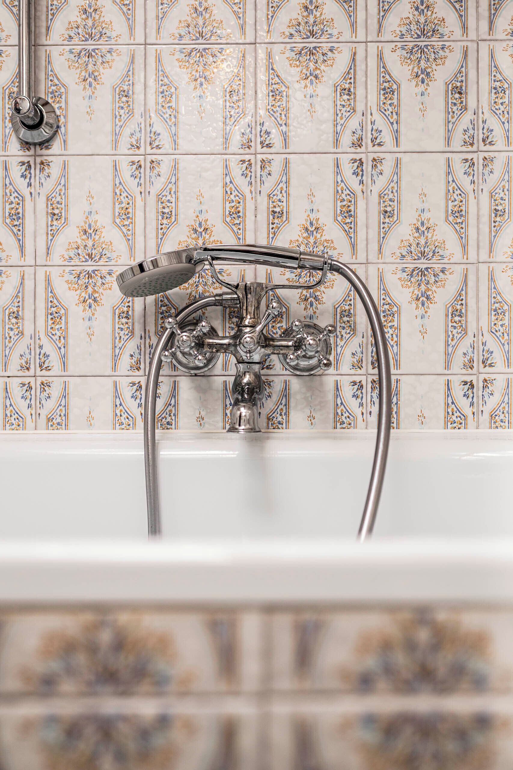 A bathroom with a white bathtub, a showerhead, and a faucet. The wall is tiled with a floral pattern.