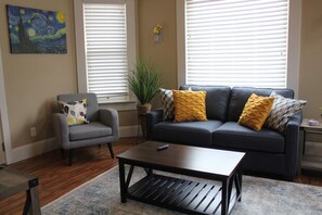 A living room with a dark sofa, a gray armchair, a coffee table, and a painting on the wall. - 131 Cedar 3 · Charming 2BR Luxury Apartment in Downtown Kalamazoo (Kalamazoo)