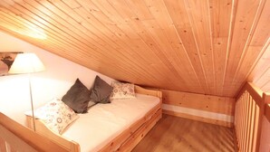 Een kleine, houten kamer met een schuine kap, een bed met kussens en een lamp.