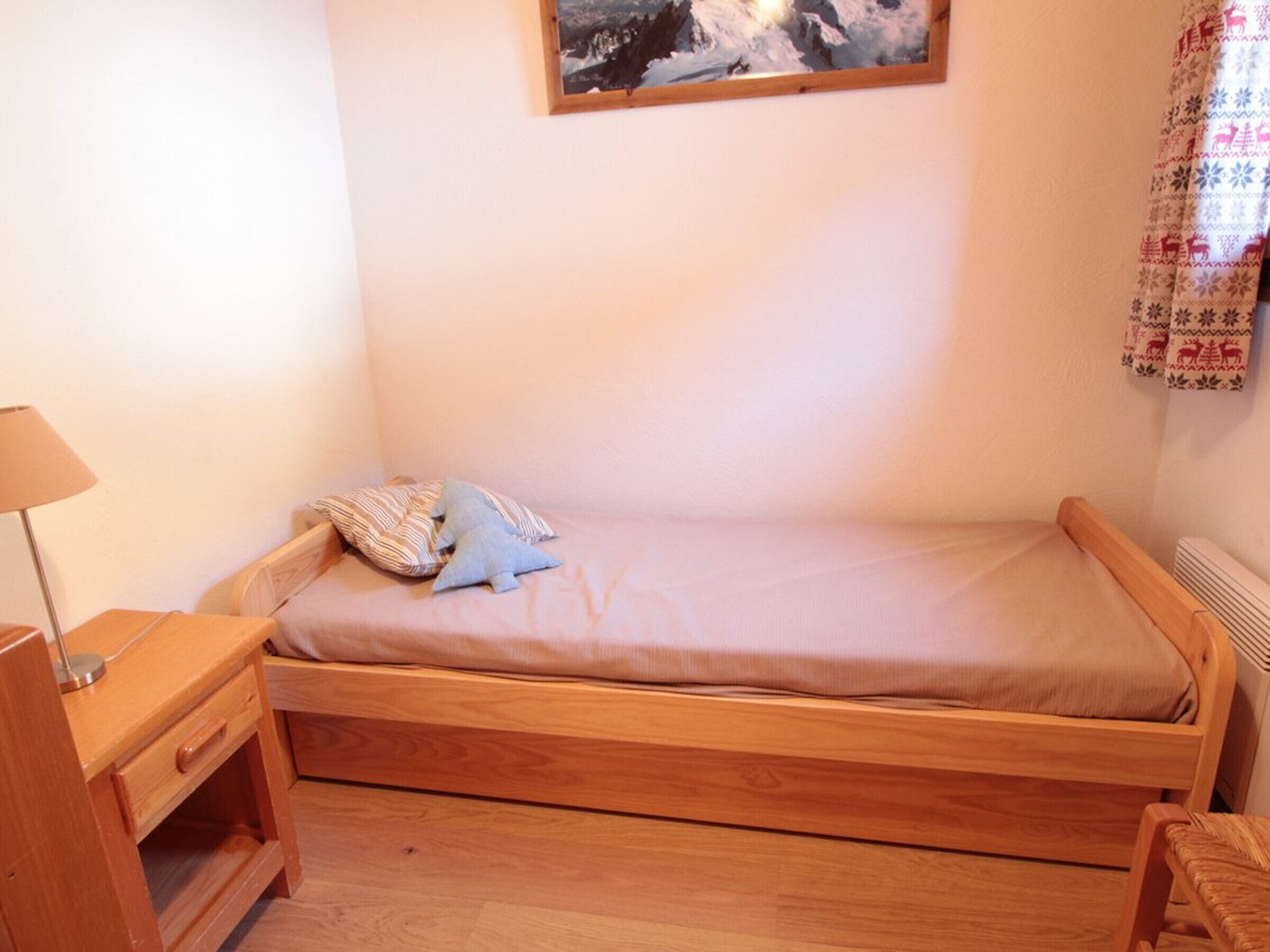 Une petite chambre avec un lit simple, un cadre en bois, une table de chevet avec une lampe et un rideau à motifs.
