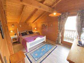Une chambre confortable dans un chalet en bois, avec un lit, des tables de chevet, un tapis bleu à motifs et un balcon agrémenté de rideaux à motifs.