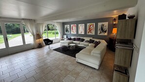 A modern living room with a sectional sofa, a coffee table, and a lamp. There are large windows with curtains, a fireplace, and artwork on the wall. - Magnifique Villa de 14 Pers Piscine Chauffée, Jacuzzi, Sauna (Saint-Martin-des-Champs)