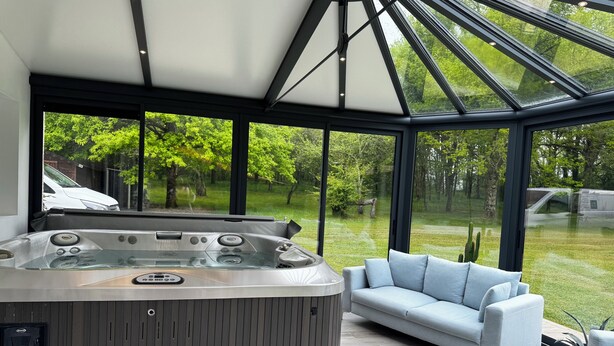 A modern spa area with a hot tub, a sofa, and a large glass window offering a view of a green garden. - Magnifique Villa de 14 Pers Piscine Chauffée, Jacuzzi, Sauna (Saint-Martin-des-Champs)