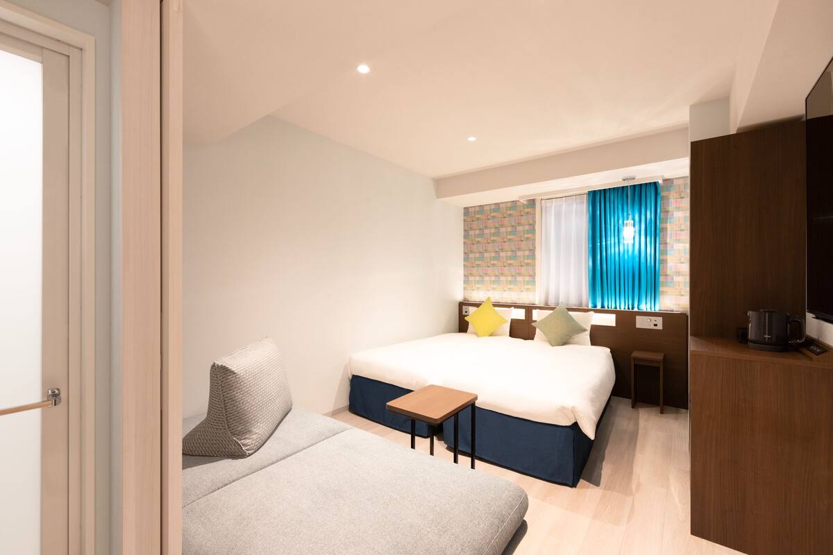 A hotel room with a bed, a sofa, a small table, a TV, and a window with curtains.
