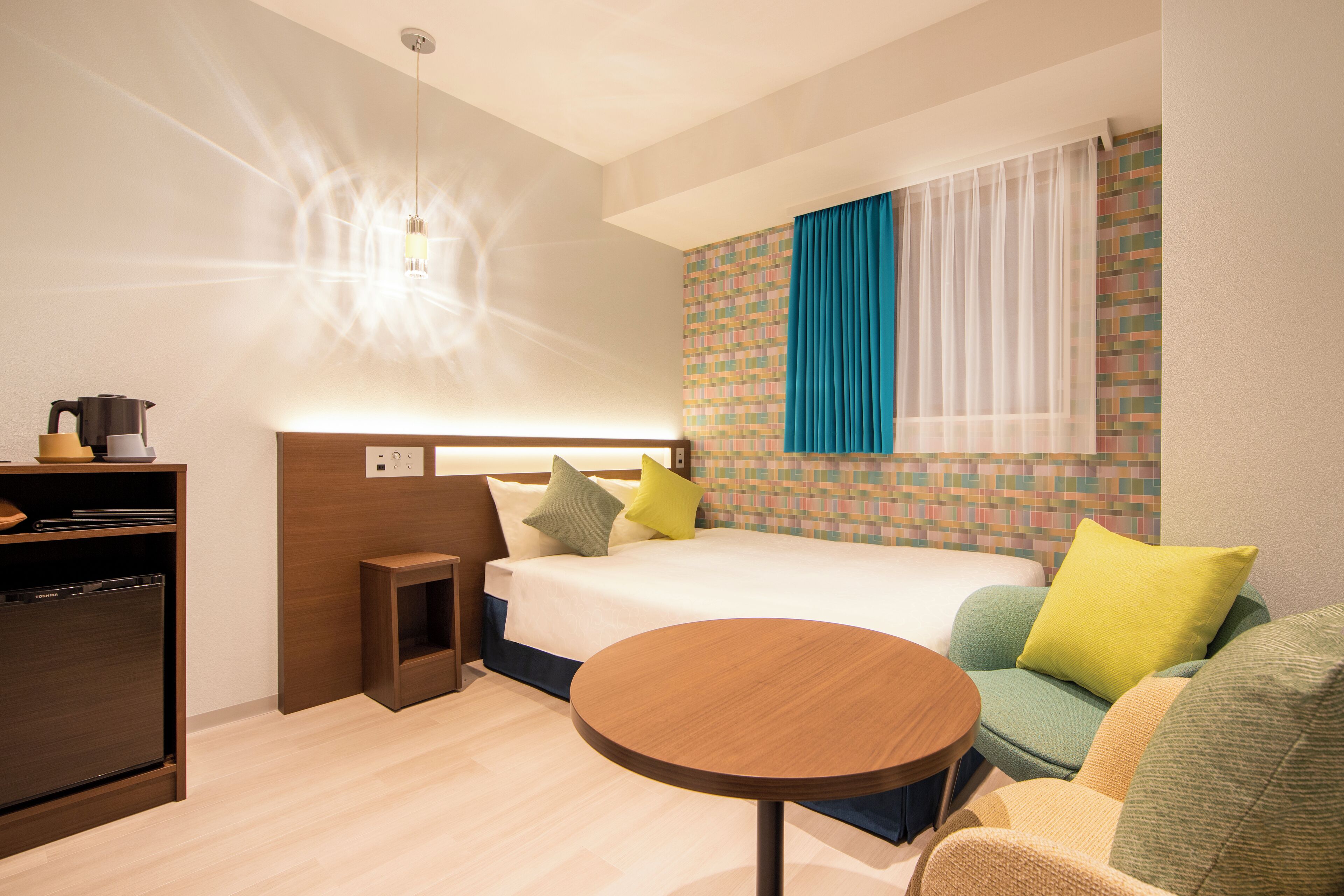 A modern hotel room with a bed, a small table, a sofa, and a kitchenette.