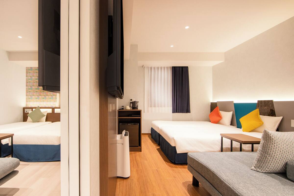 A hotel room with two beds, a sofa, a TV, a desk with a computer, and a window with curtains.