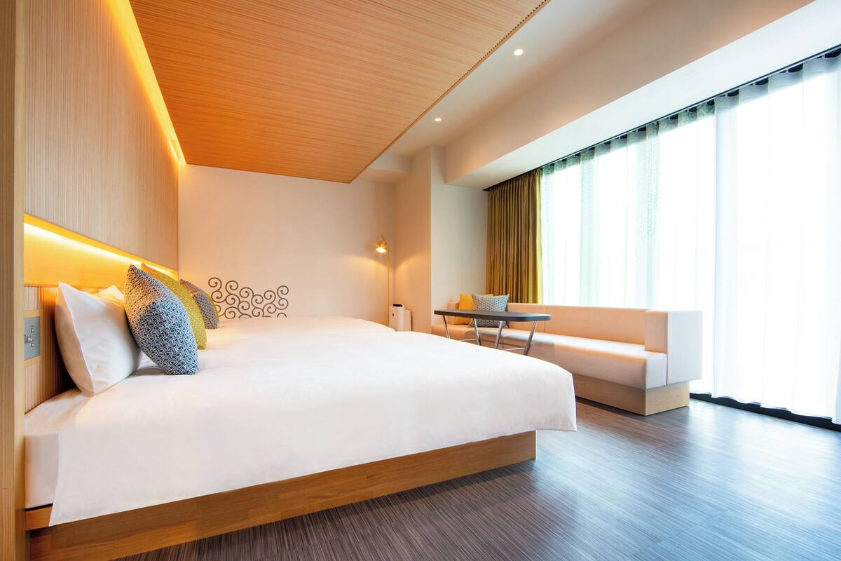 A modern hotel room with a large bed, a small table, a sofa, and a large window with curtains.