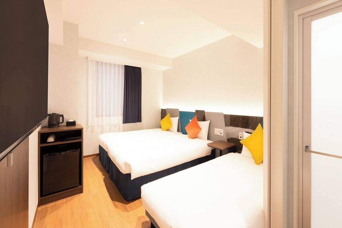 A hotel room with two beds, a desk, a coffee machine, and a window with curtains.