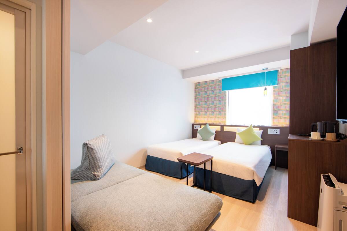 A hotel room with two beds, a desk, a chair, a TV, and a coffee machine.
