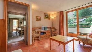 Een knusse woonkamer met een houten tafel, rieten stoelen, een televisie op een houten kast en een groot raam met uitzicht op groen.