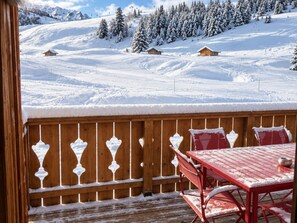 Une terrasse en bois avec une table rouge et des bancs, d’où l’on peut admirer un paysage montagneux enneigé parsemé de maisons.