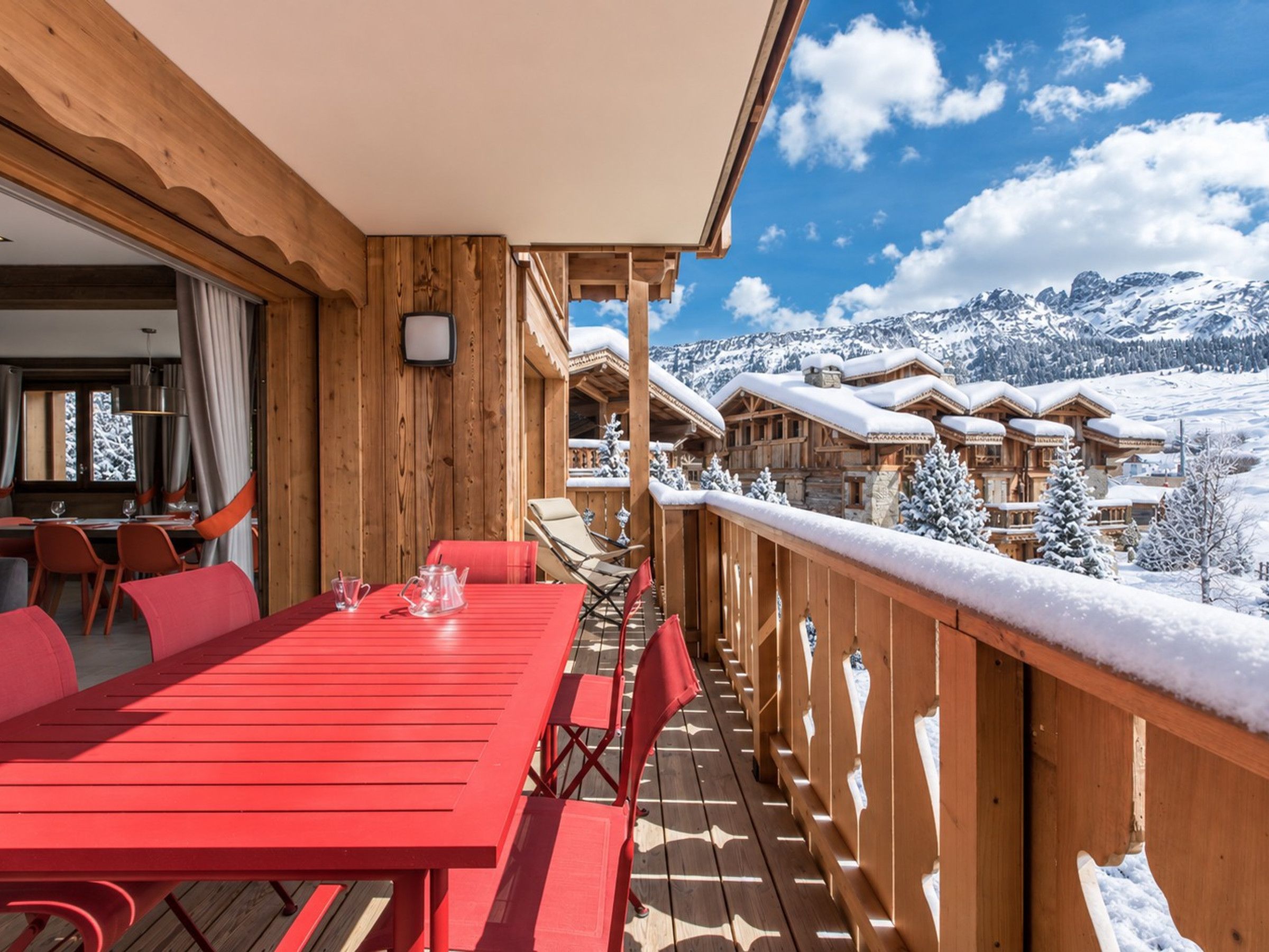 A wooden deck with a red table and chairs, overlooking a snowy mountain landscape.