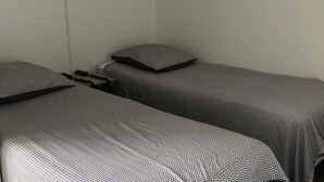 Une petite chambre, de taille modeste, avec deux lits simples, chacun recouvert d’une couvre-lit à motifs. On y trouve une table de chevet avec une lampe et une prise électrique au mur.
