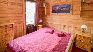 Une pièce de style cabane en bois, avec un lit, une fenêtre avec des rideaux, un tableau accroché au mur et une petite commode en bois.