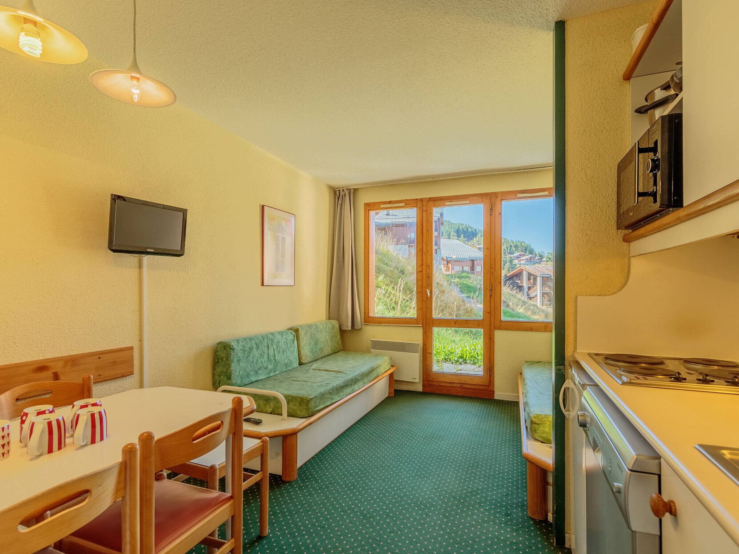 A compact kitchen with a dining area, a green sofa, and a view of houses through the window.