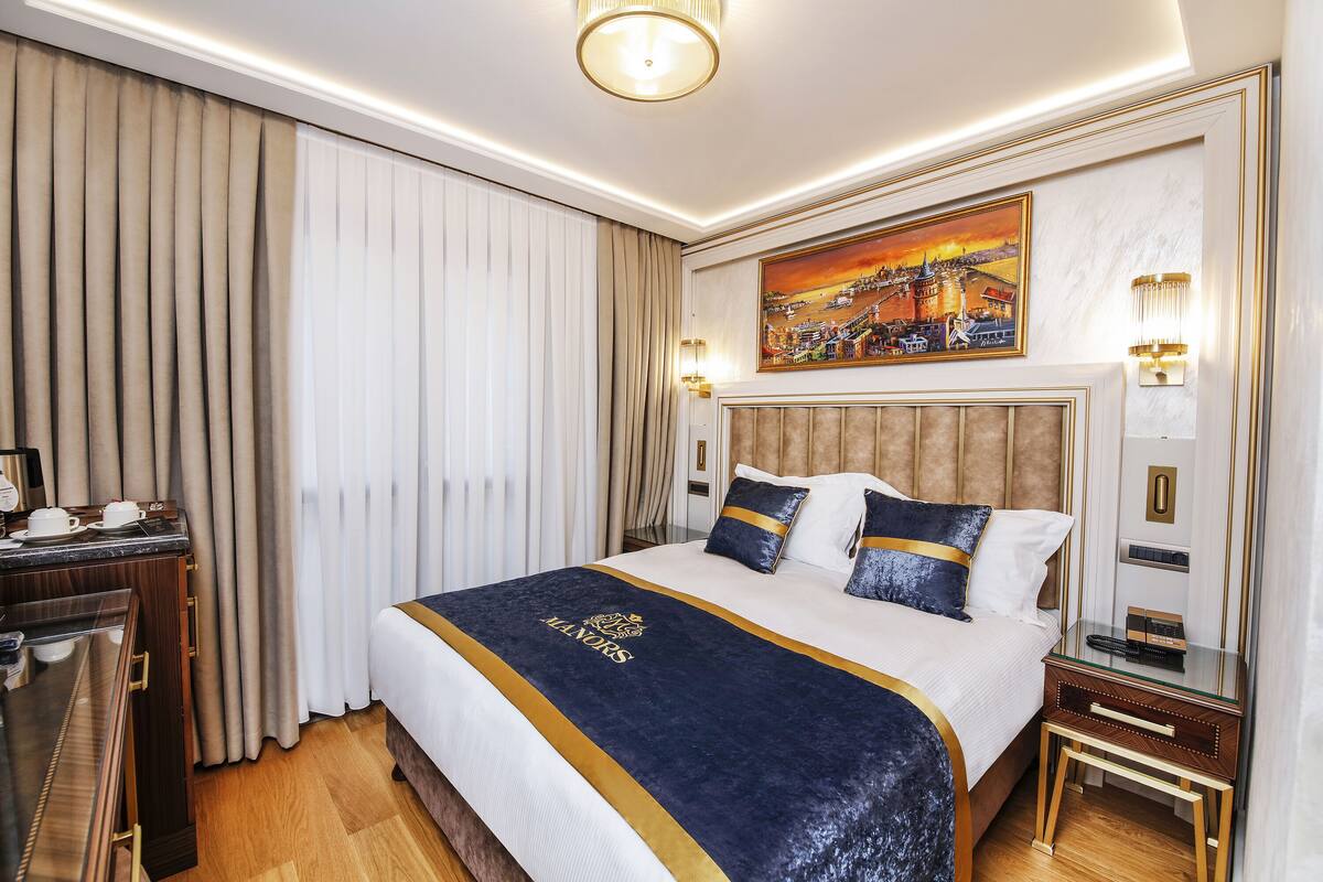 A hotel room with a large bed, a bedside table, a desk with a chair, and a painting on the wall.