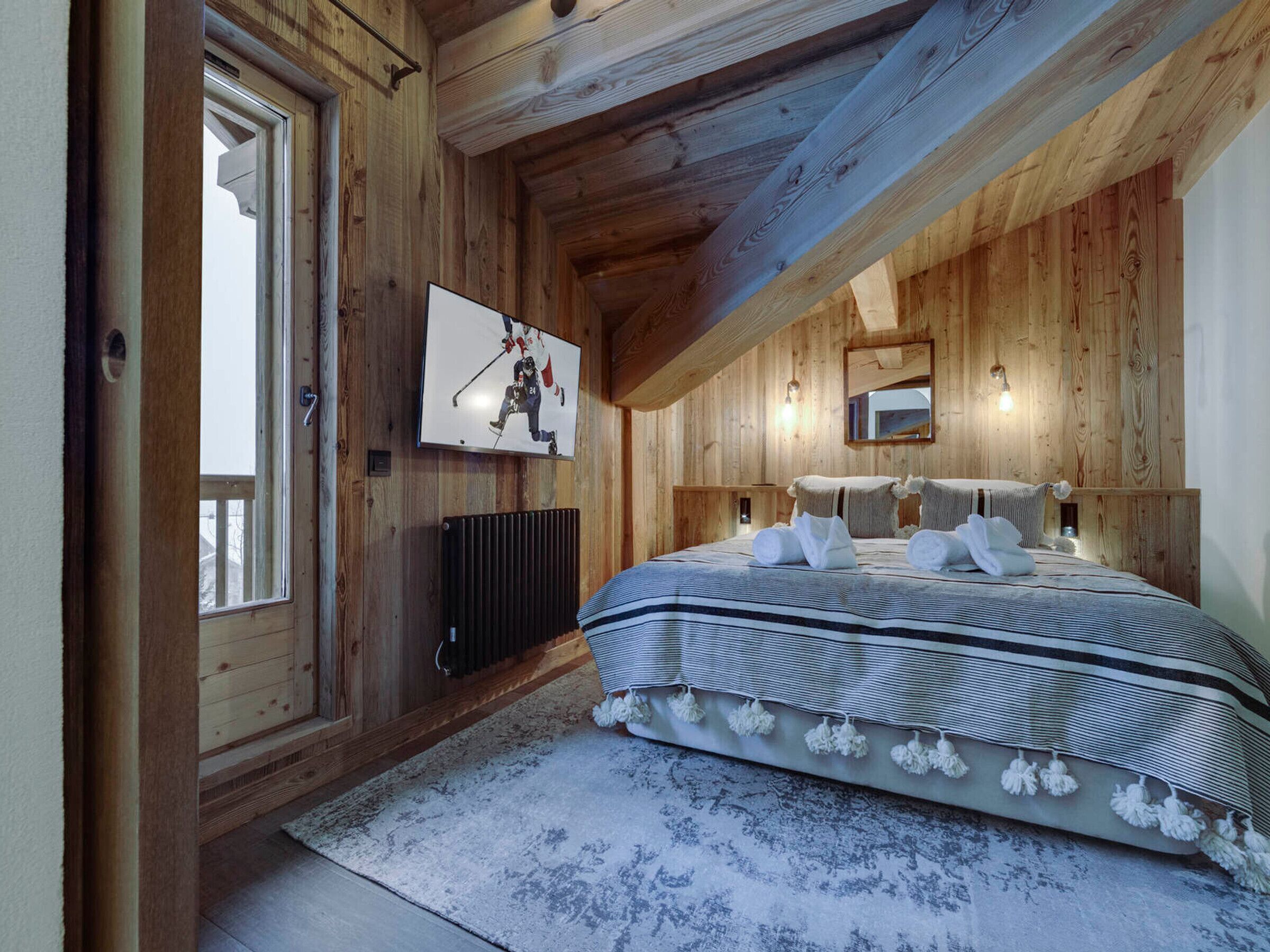 Un dormitorio con paneles de madera, una cama, un televisor montado y un ventanal.