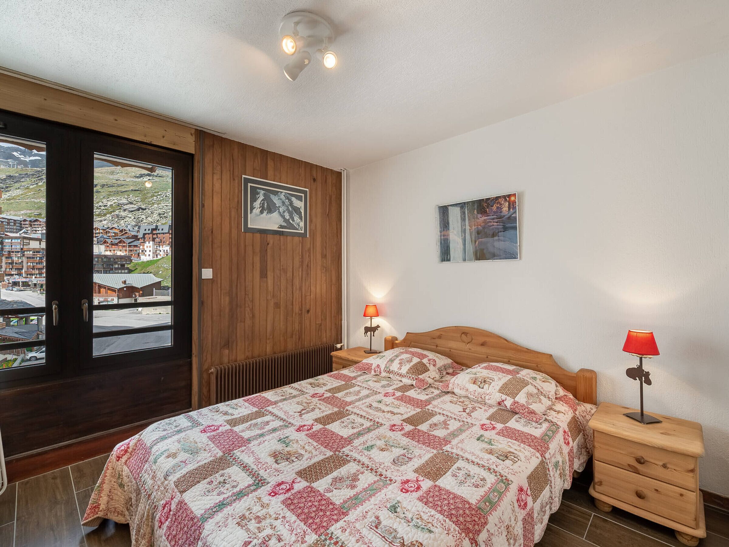 A bedroom with a wooden bed, a nightstand, a red lamp, a framed picture on the wall, and a view of a mountainous area through a door.