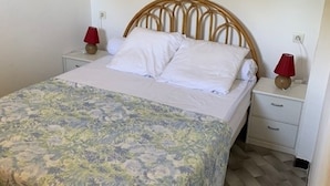 Un lit avec une tête de lit en bois, une literie blanche et une couvre-lit à motifs floraux. Il y a deux tables de chevet avec des lampes et un petit tiroir.