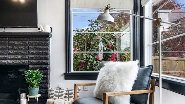Un coin douillet avec une cheminée en pierre, un fauteuil confortable agrémenté d’un coussin moelleux, une petite table avec une plante et une fenêtre donnant sur un paysage verdoyant, ainsi qu’une lampe.
