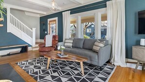 Un salon avec un canapé, une table basse et un fauteuil. On aperçoit un escalier au fond.