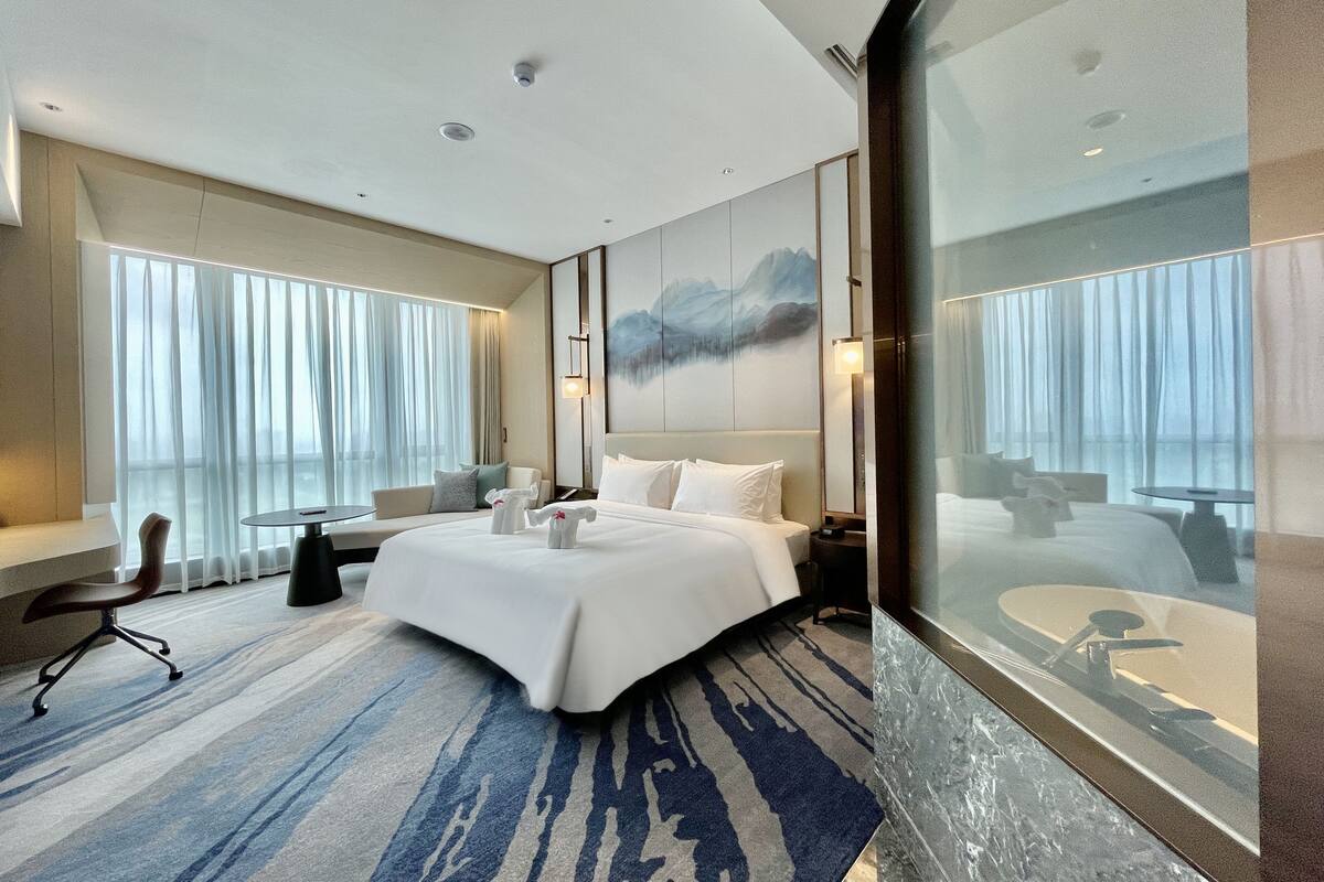 Habitación de hotel moderna con una cama grande, un escritorio y vistas a través de un muro de cristal.