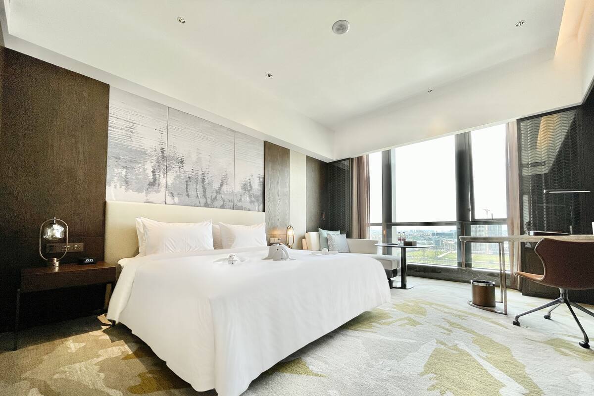 Una habitación de hotel moderna con una cama grande, un escritorio y vistas de la ciudad a través de amplios ventanales.