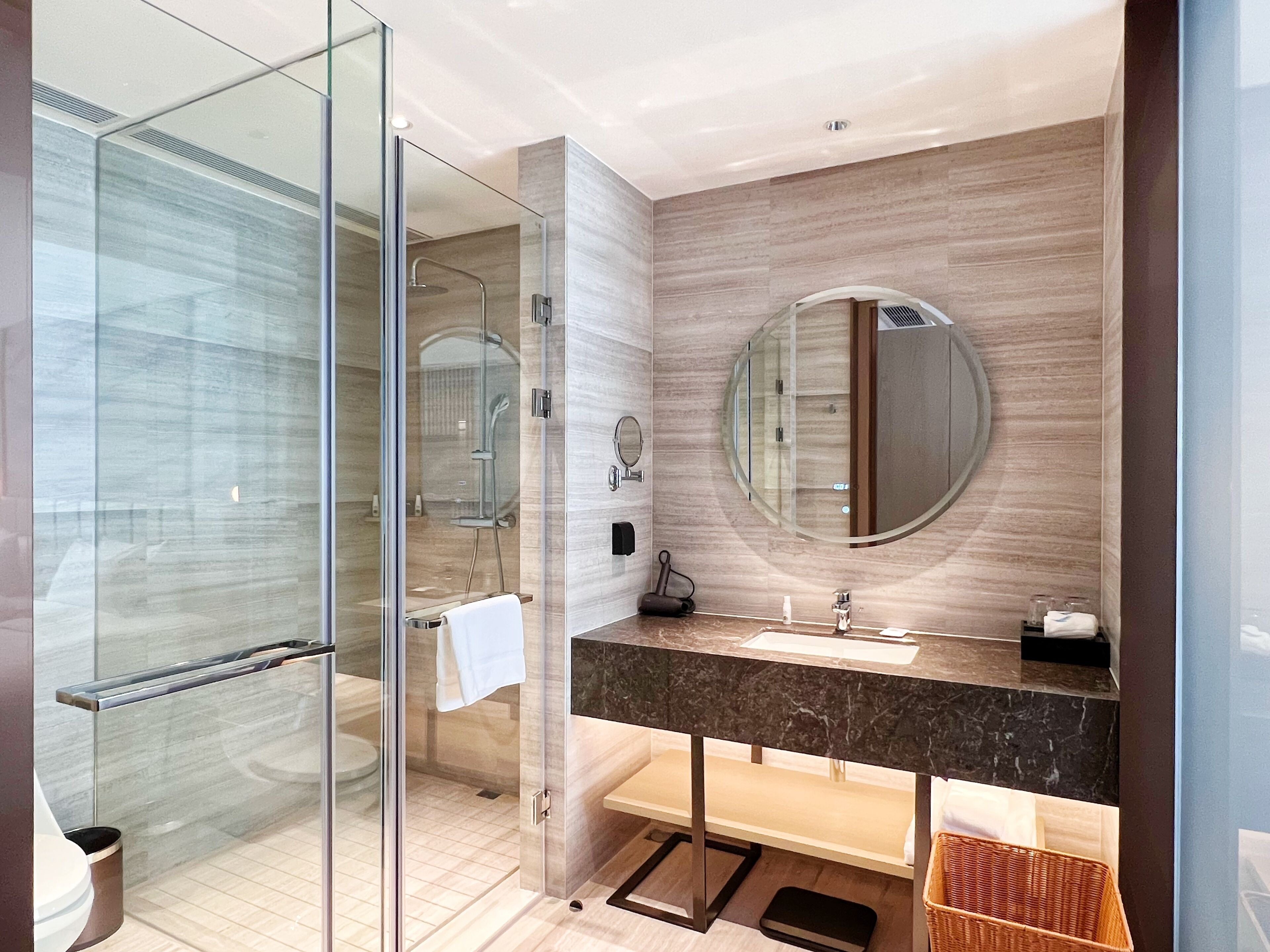 A modern bathroom with a glass shower enclosure, a round mirror, and a marble countertop.