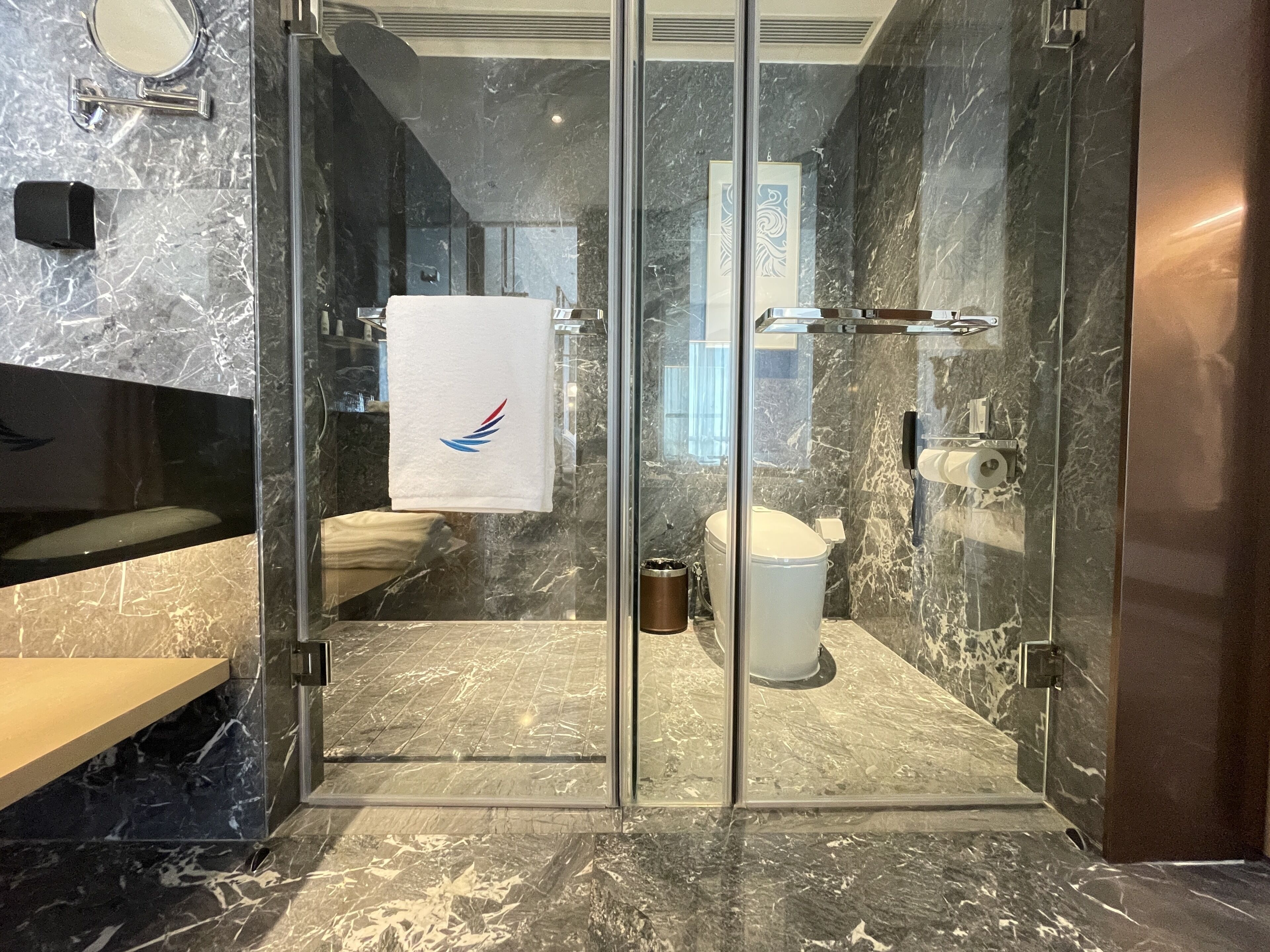 Een moderne badkamer met een glazen douchecabine, marmeren vloer, toilet en een wastafel met een handdoek aan de muur.