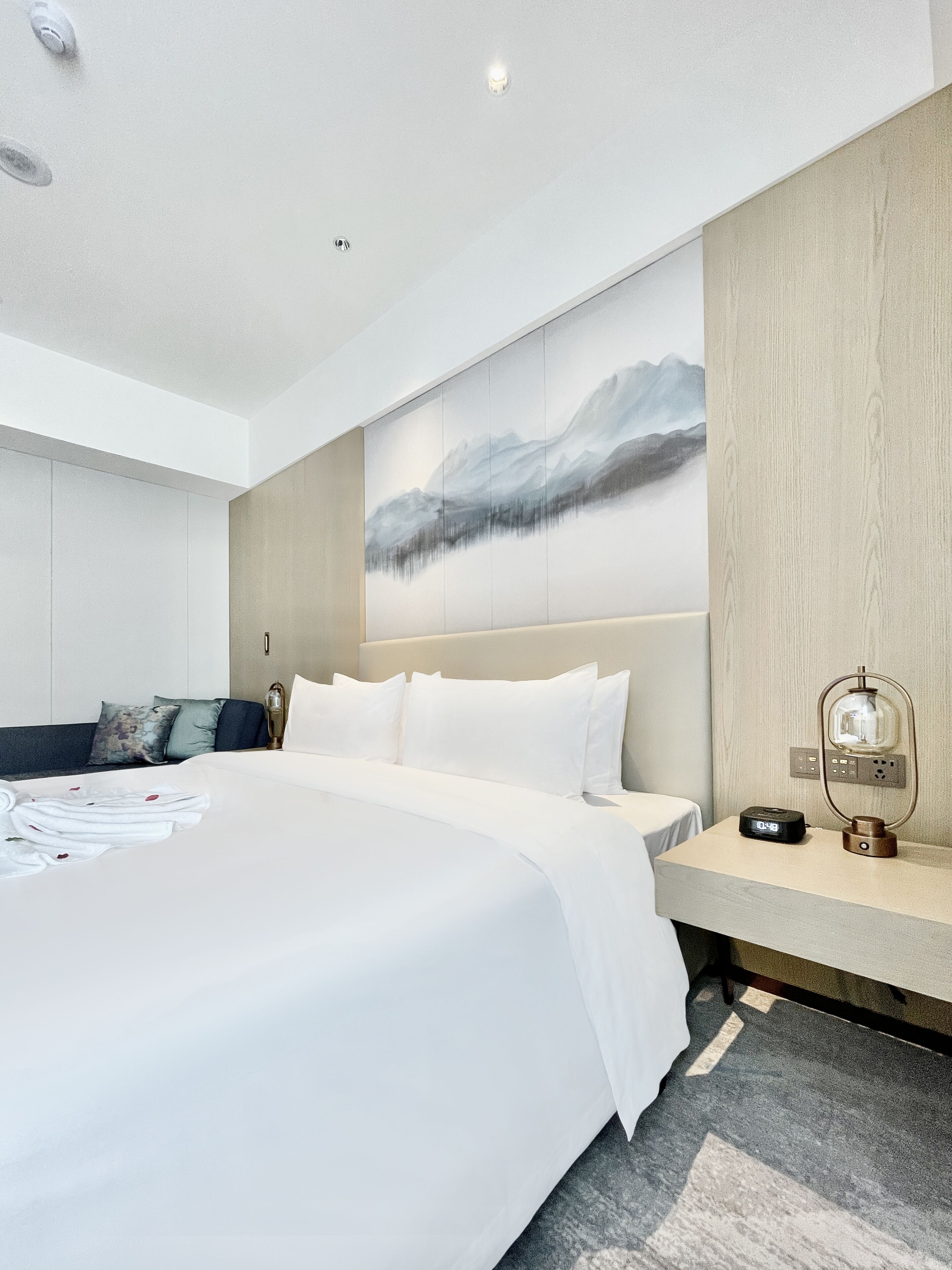 A modern hotel room with a large bed, a nightstand with a lamp, and a painting of mountains on the wall.