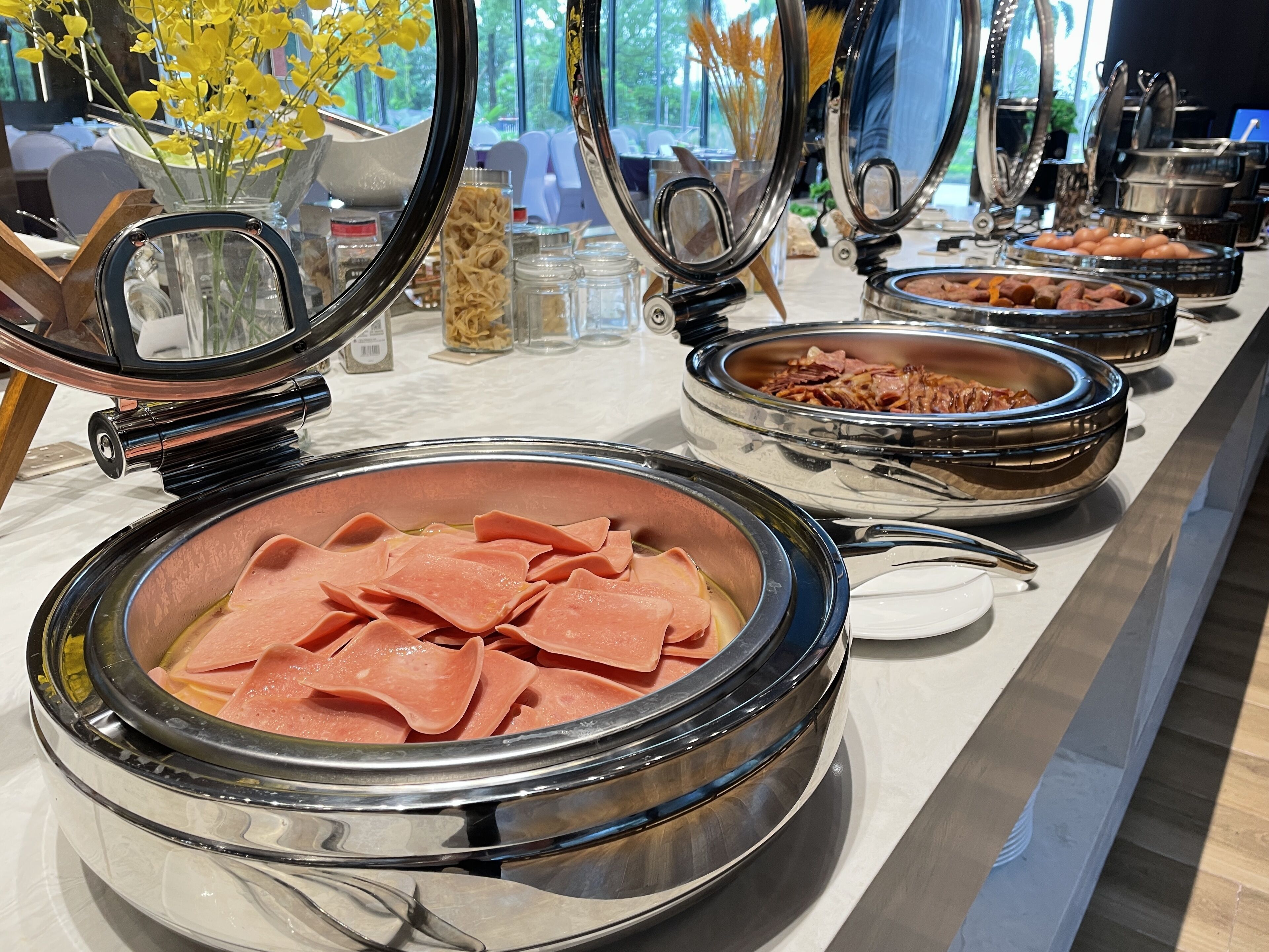 Un bufé con diversos alimentos, incluyendo carnes y verduras en porciones, dispuesto en un mostrador con vistas al exterior.