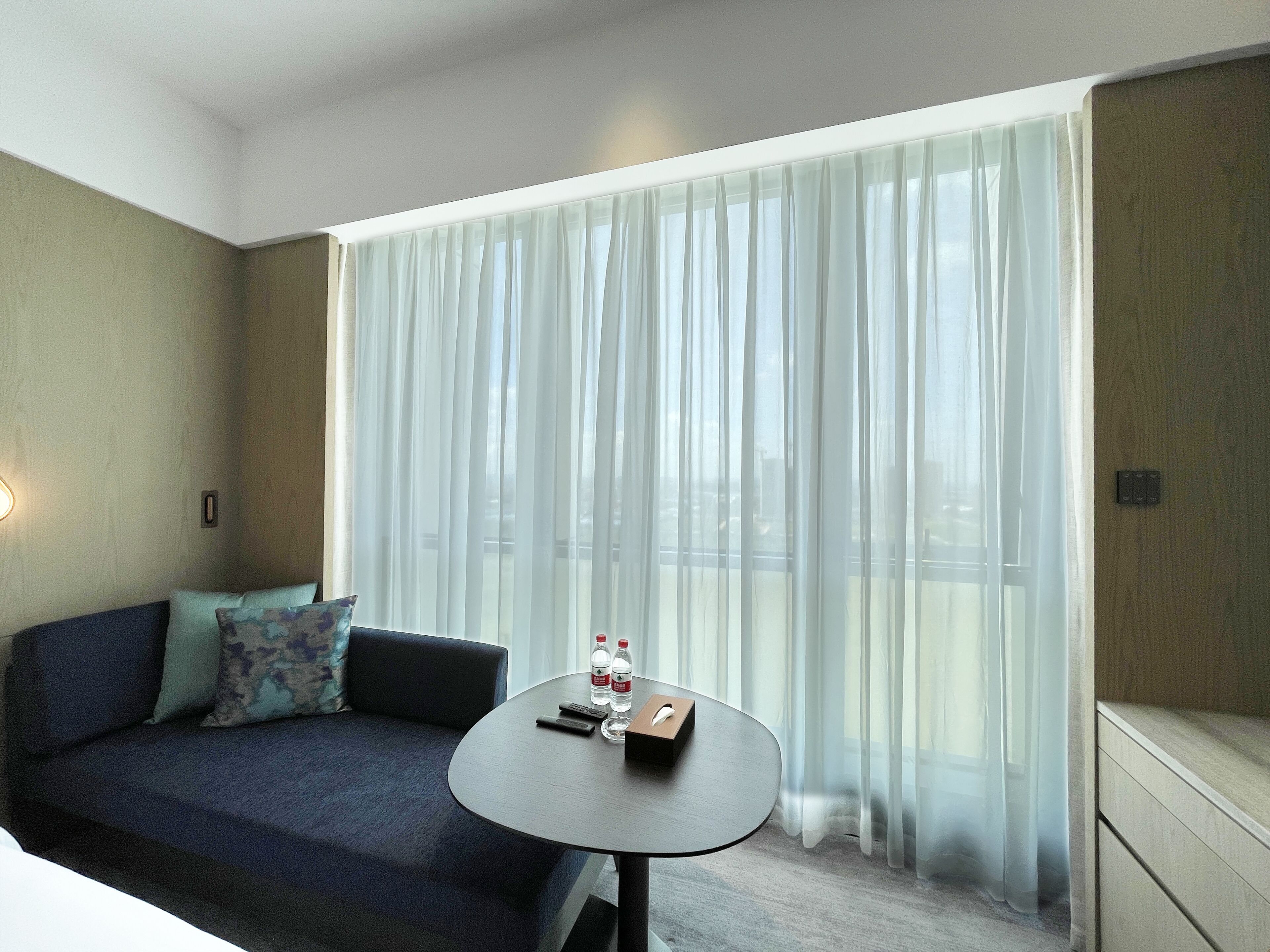 A hotel room with a dark blue sofa, a small round table with a remote control, a bottle of water, and a tissue box.