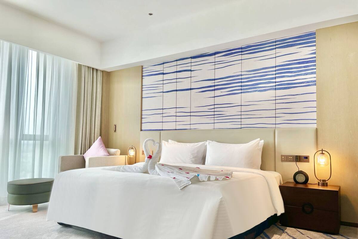 Una habitación de hotel con una cama grande, una mesita de noche con lámpara, una obra de arte en la pared y una ventana con cortinas traslúcidas.