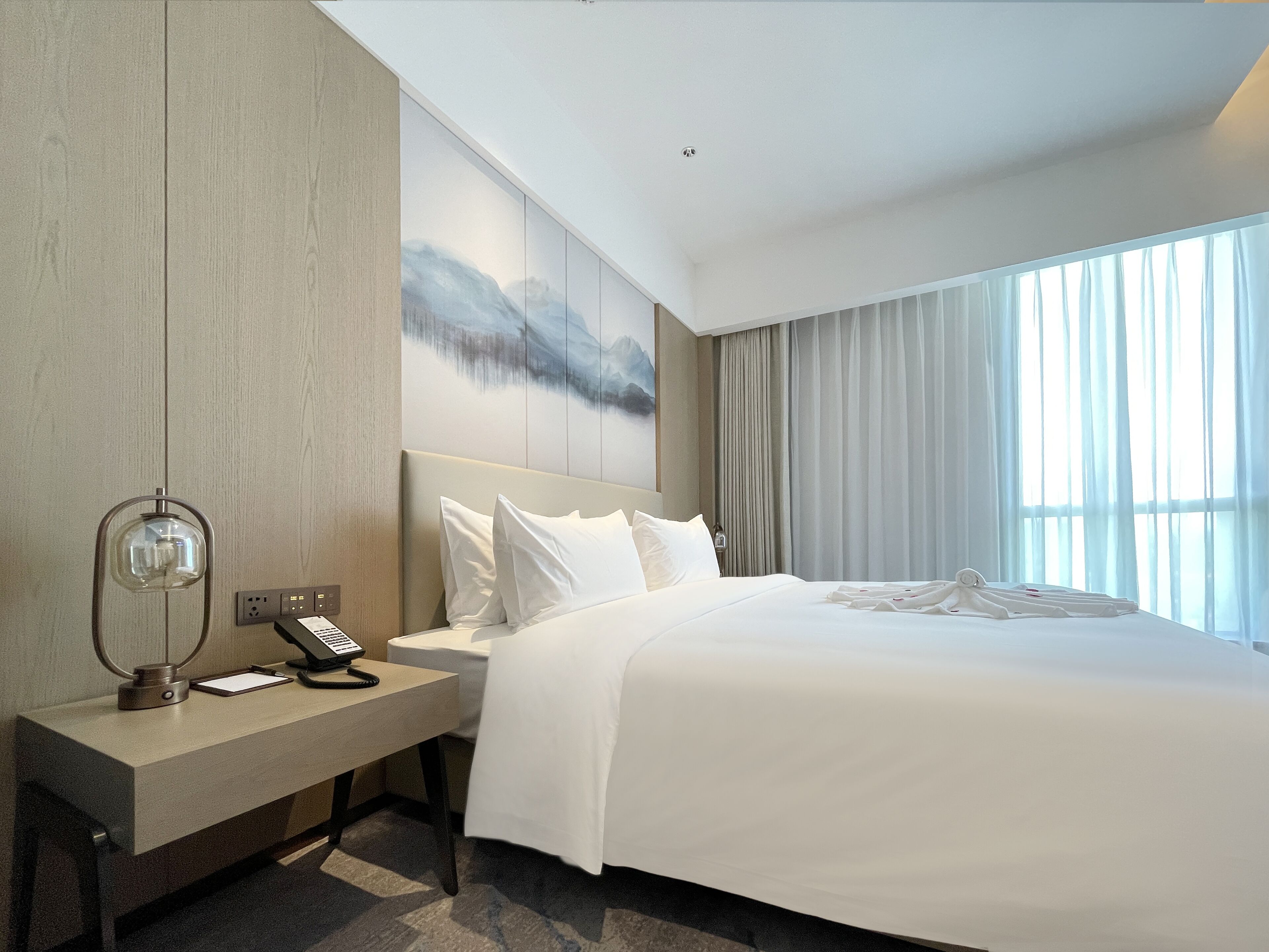 A modern hotel room with a large bed, a bedside table with a lamp, and a painting on the wall.