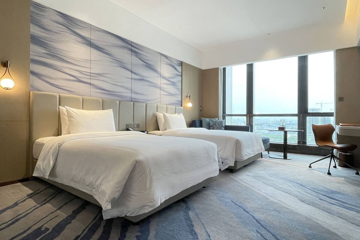 Habitación de hotel con dos camas, un ventanal amplio con vista a la ciudad, una mesita pequeña y una silla.