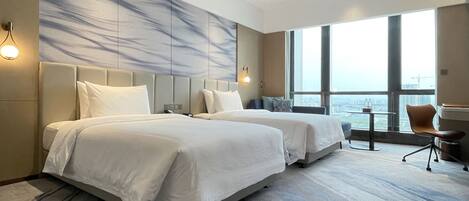 A hotel room with two beds, a large window with a city view, a small table, and a chair.
