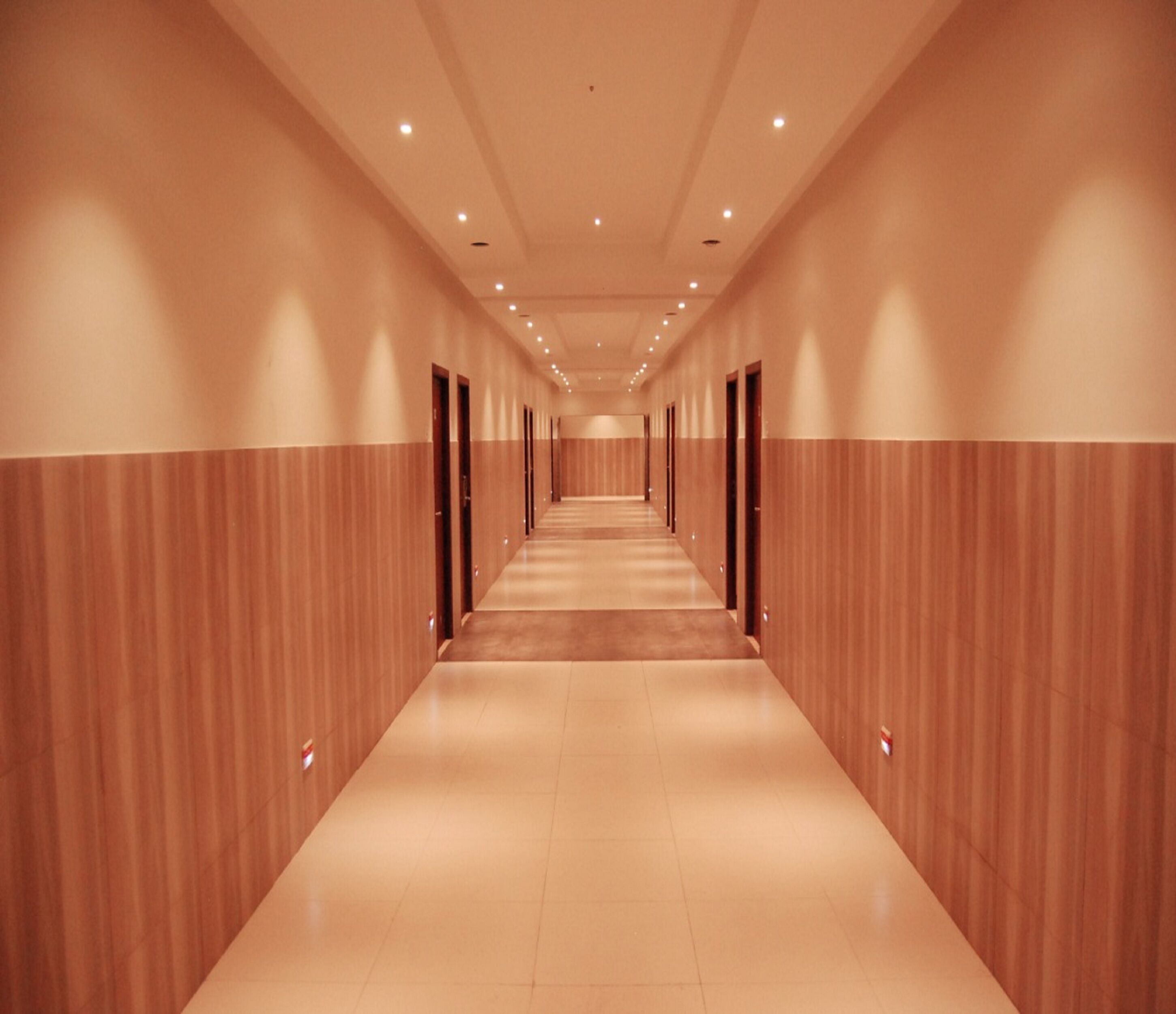 Un largo pasillo bien iluminado con paneles de madera en las paredes y piso de baldosas, que conduce a varias puertas.