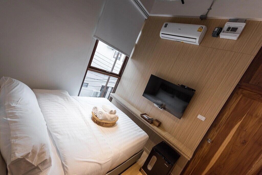 A hotel room with a bed, a TV mounted on the wall, an air conditioning unit, and a window providing natural light.